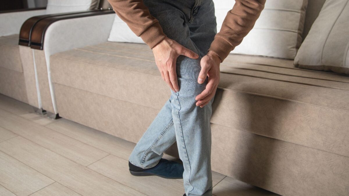Knee pain when standing up from sitting treatment - Spine and Joint Pain Center Person sits on a beige sofa, clutching their knee with both hands, suggesting knee pain or injury.