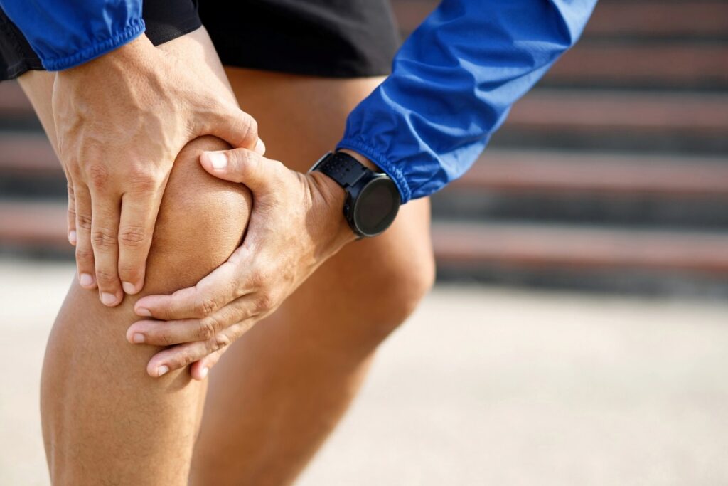 common causes of knee pain when standing from sitting position - Spine and Joint Pain Center Person in a blue jacket grasps their knee with both hands, indicating knee pain or injury.